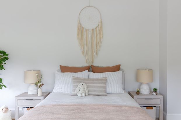A bright, calm bedroom viewed straight on, featuring a neatly made bed with white striped bedding, layered rust and neutral pillows, a soft beige throw, matching white bedside tables and lamps, small plants, and a woven hanging wall decoration above the bed. The room has white walls and a simple, symmetrical contemporary arrangement.
