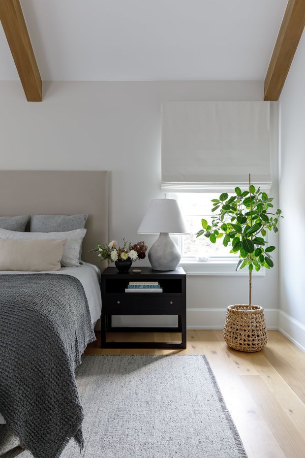 A bright, softly styled bedroom with a neutral upholstered bedhead, layered grey and beige bedding, a black bedside table, and a sculptural ceramic lamp. Light timber flooring, a pale textured rug, white walls, exposed timber ceiling beams, and a roller blind frame a sunlit window with a potted indoor tree in a woven basket.