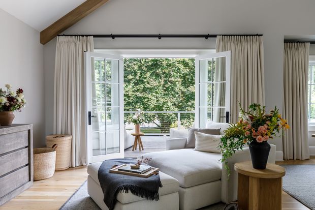Bright, refined living room with a large white sectional sofa and ottoman, layered neutral textiles, pale timber flooring, full-height cream curtains, and French doors opening to a balcony or terrace. A substantial exposed timber ceiling beam adds architectural character, while greenery and floral arrangements bring natural colour indoors.