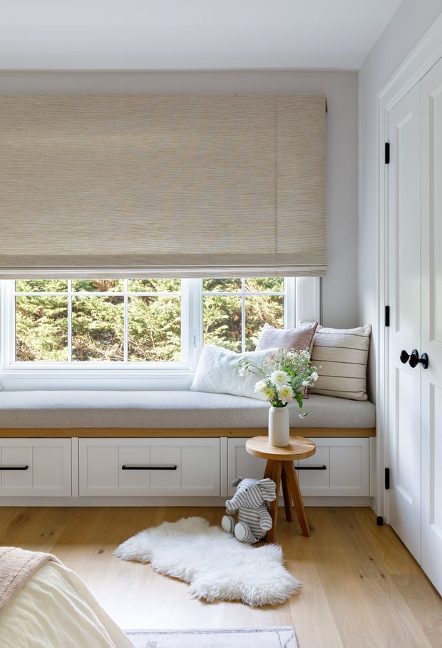Bright, serene bedroom corner featuring a built-in window seat with white cabinetry and a light timber bench top, large divided-light windows, a woven Roman blind, pale walls, and a white paneled door. Soft cushions, a small timber side table, plush sheepskin rug, and timber flooring create a warm, refined retreat.