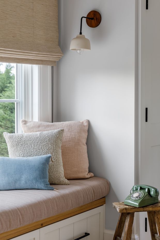 A bright, compact window seat nook with a built-in upholstered bench, white paneled cabinetry, and layered textured cushions in blush, cream, and blue. A woven roman blind frames the window, while a wall-mounted lamp with a timber backplate provides focused lighting. A vintage green telephone sits on a rustic wooden stool beside the seat. The palette is soft, warm, and inviting with natural timber accents.