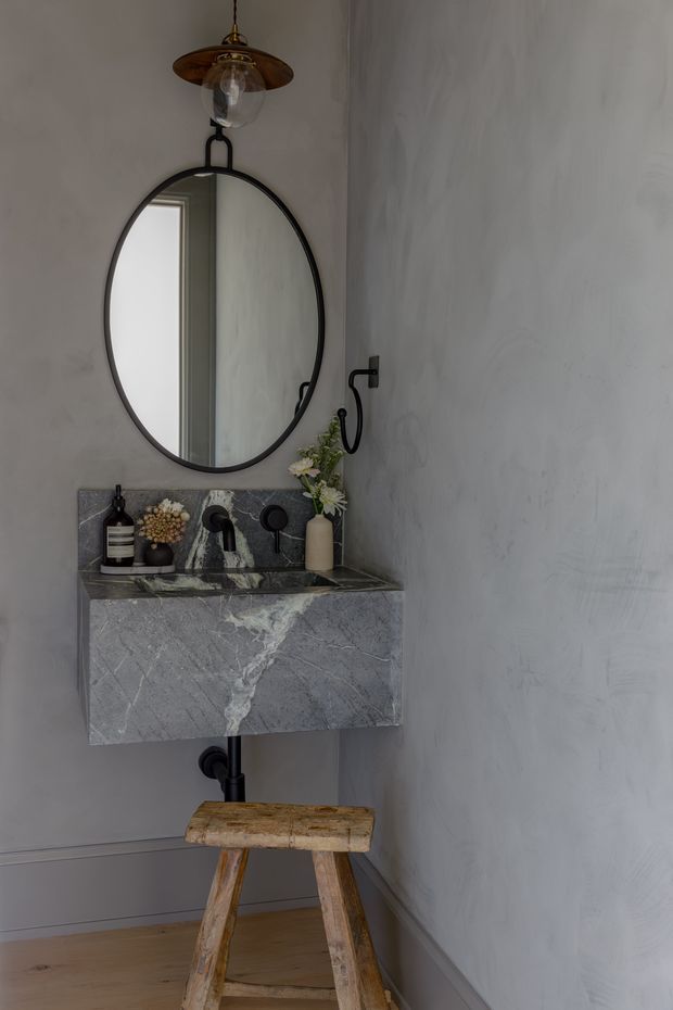 A compact, moody powder room with a wall-mounted grey veined stone basin and integrated backsplash, matte black tapware and exposed plumbing, a large oval black-framed mirror, and a vintage brass pendant light. Textured pale grey walls and timber flooring create a refined, rustic character, with a small weathered wooden stool beneath the basin.