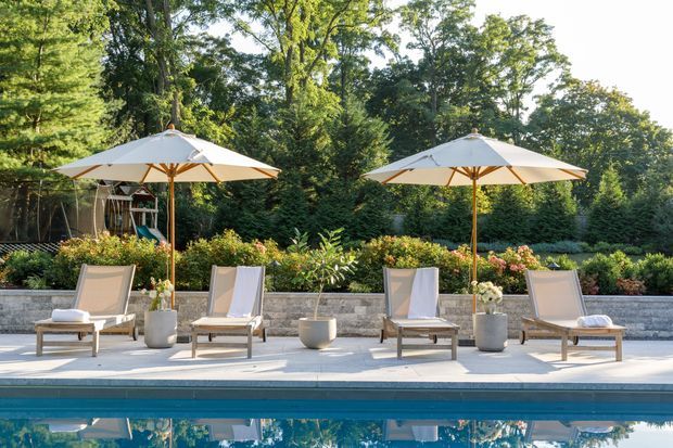 A landscaped outdoor pool area with a calm, symmetrical arrangement of four cushioned sun loungers beneath two large white umbrellas. The loungers sit on a pale paved terrace beside a turquoise swimming pool, with large planters, flowering garden beds, a low stone retaining wall, and dense mature trees forming a lush backdrop.
