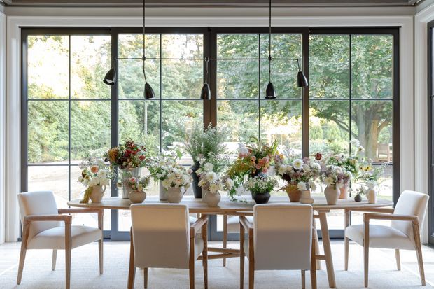 Bright, elegant dining area centered on a long light-wood dining table surrounded by upholstered dining chairs. Large black-framed steel-look windows provide extensive views into lush greenery, while multiple sculptural pendant lights hang above a floral-filled tabletop. The room has pale walls, a light floor covering, and a refined indoor-outdoor connection.