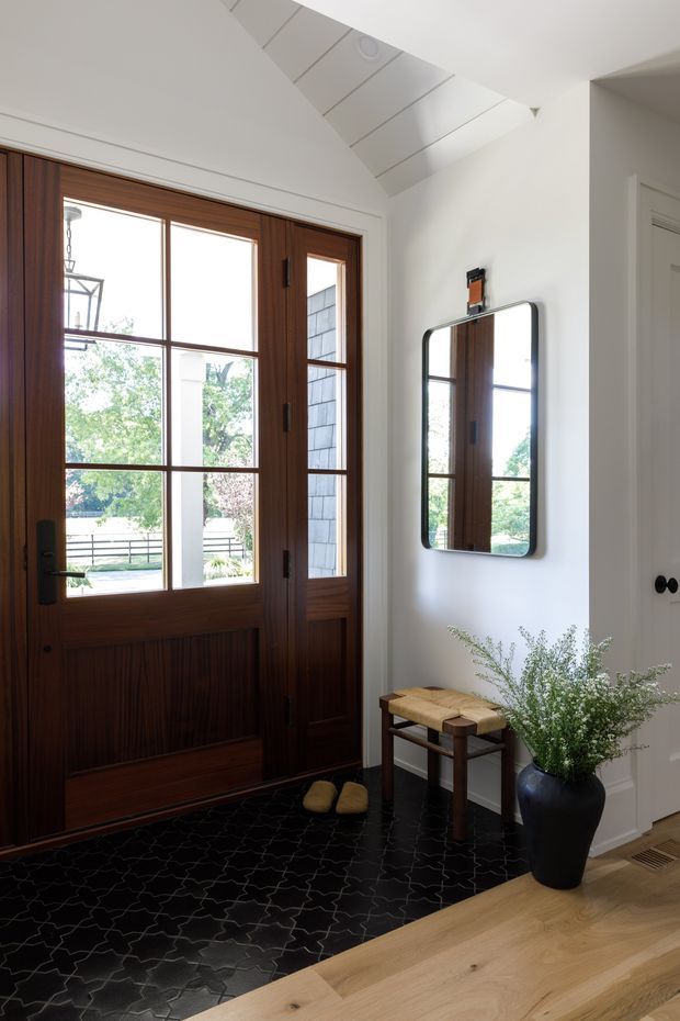 A refined residential entryway with a dark stained timber glazed front door and matching sidelights, white walls and trim, a black patterned tile floor, and a warm light-oak timber floor beyond the tiled threshold. A rounded-rectangle wall mirror, small wooden bench, and potted greenery sit beside the door. The space has a bright vaulted ceiling with linear panel detailing and a skylight-like glazed section.