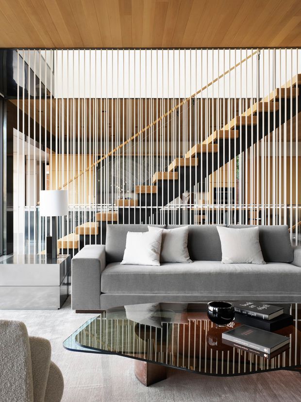 A sophisticated contemporary living room with a large grey upholstered sofa, white cushions, a glossy black glass coffee table, and a pale textured rug. Behind the seating area is a dramatic open staircase with dark stringers, timber treads, and a timber handrail, screened by vertical white and timber slats. The ceiling is clad in warm timber boards, and extensive glazing and reflective surfaces create a bright, layered interior.