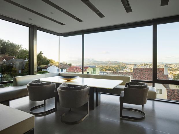 A contemporary dining area with a large white rectangular table, upholstered cantilever chairs, and built-in bench seating positioned beside expansive corner windows. The room has a smooth grey floor, dark window frames, a white ceiling with recessed strip lighting, and broad views over rooftops and rolling hills.