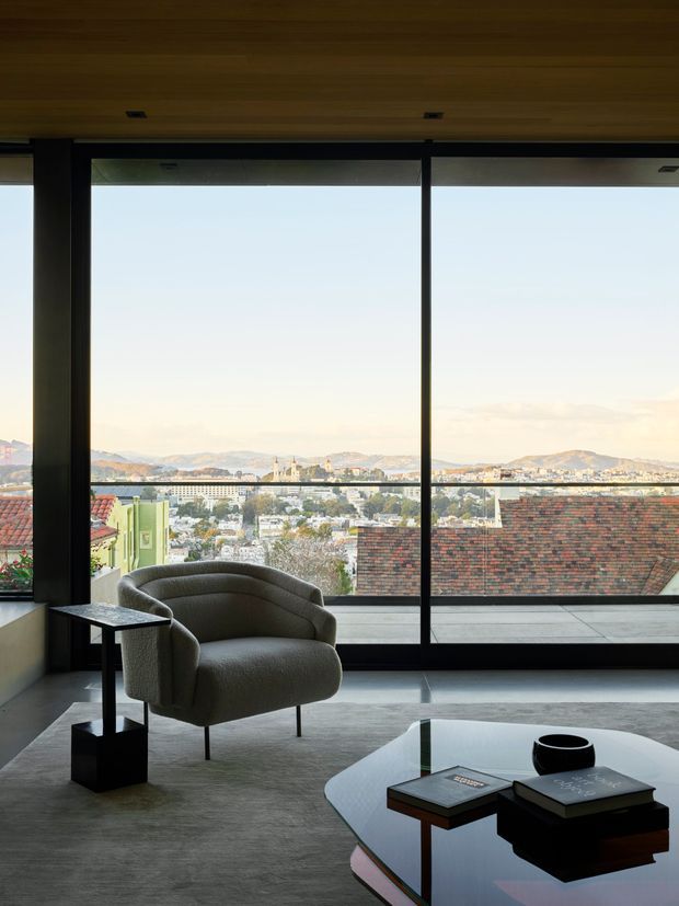 A refined living room with expansive floor-to-ceiling glazing overlooking a dense cityscape and hills. The room features a sculptural upholstered lounge chair, small black side table, low glossy coffee table, and a broad grey rug. Warm timber ceiling panelling contrasts with dark window frames and a restrained neutral palette.