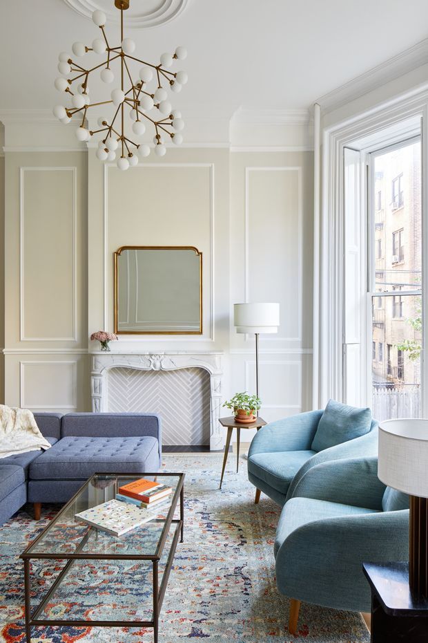 Elegant, bright living room with refined traditional architectural detailing. The room features pale moulded wall panelling, decorative ceiling medallion and cornices, an ornate white fireplace with herringbone tile surround, large sash-style window, contemporary brass-and-white globe chandelier, blue upholstered seating, a glass coffee table, patterned area rug, and a framed mirror above the mantel.