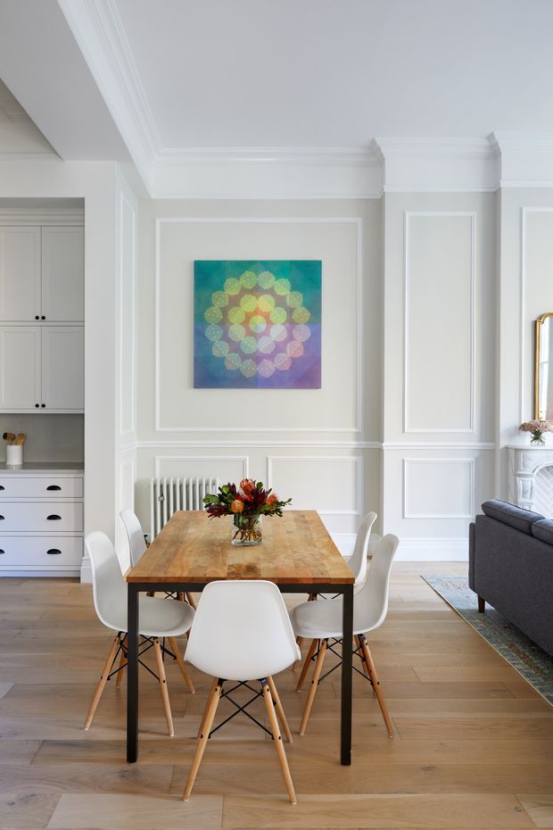 Bright, refined dining area in a classic apartment interior. A rectangular natural timber dining table with white molded chairs sits on a warm timber floor. The room features white decorative wall panelling, ornate crown moulding, built-in cabinetry, a traditional radiator, and a colourful abstract artwork. A partial living room with a dark sofa and fireplace or mirror is visible to the right.