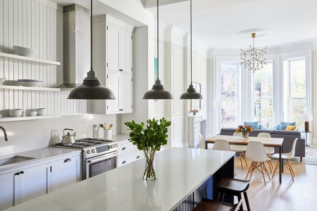 Bright, open-plan kitchen and dining area with white shaker cabinetry, a large pale stone island, vertical wall panelling, pendant lights, and a stainless-steel range. The dining space and living area extend toward tall glazed doors and windows, with ornate ceiling detailing and classic trim throughout.