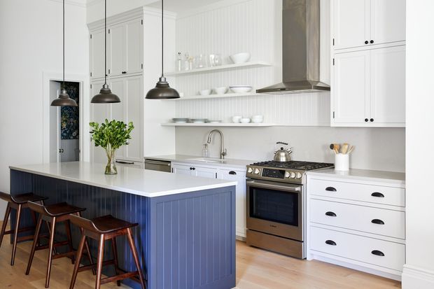Bright, refined kitchen interior with a large island featuring blue vertical-panel cabinetry and white benchtop, paired with timber stools. The room has extensive white shaker-style cabinetry, open shelving on a vertically panelled wall, a freestanding stainless-steel range cooker, aged metal rangehood, pendant lights, and pale timber flooring.