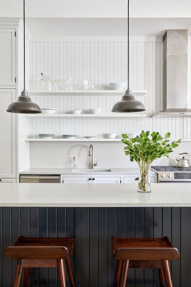 Bright, refined kitchen featuring white vertical wall panelling, open display shelves with glassware and crockery, pale stone benchtops, white cabinetry, a stainless-steel rangehood and cooker, and a dark vertically panelled island. Two large aged-metal pendant lights hang above the island, with mid-tone timber bar stools and a vase of greenery adding contrast.