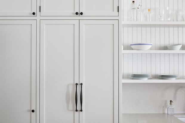 Close-up of a bright white built-in kitchen or butler's pantry, featuring shaker-style cabinetry with recessed panel doors and slim black hardware. Open white shelving displays glassware, bowls and stacked plates, while a pale stone-look benchtop and subtle white textured backsplash are visible at the lower right.