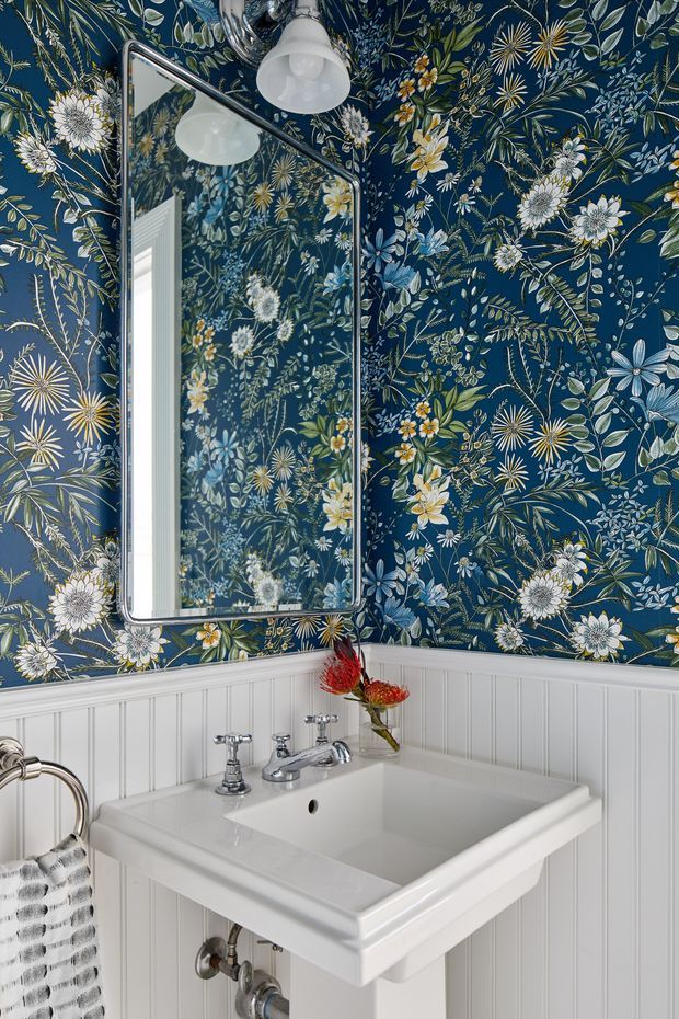 A compact bathroom or powder room featuring a white pedestal-style wall-mounted basin with polished chrome taps, exposed plumbing, a rectangular chrome-framed mirror, and white vertical beadboard wall panelling. The upper walls are covered in a dramatic navy floral botanical wallpaper with dense white, blue, green, yellow, and muted orange flowers and foliage. A small floral arrangement adds colour beside the basin, and a striped hand towel hangs at the left.