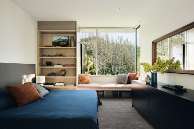 A refined bedroom with a deep blue upholstered bed in the foreground, warm timber built-in shelving, a full-width window seat, and expansive glazing overlooking a wooded landscape. A dark contemporary dresser with a large timber-framed mirror occupies the right side, while layered cushions, books, ceramics, and greenery add warmth. The room is bright, calm, and contemporary with a strong connection to nature.