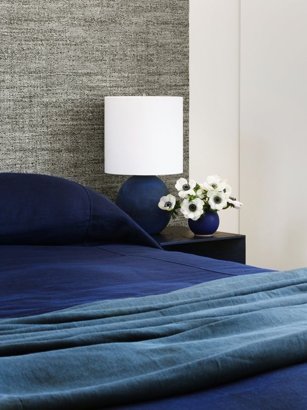 Close-up of a stylish bedroom with deep navy bedding and pillows, a pale blue folded throw across the bed, and a dark bedside table holding a white-shaded lamp and blue ceramic vases with white flowers. The backdrop features a textured grey fabric wall panel beside a smooth off-white wall, creating a refined, tonal interior.
