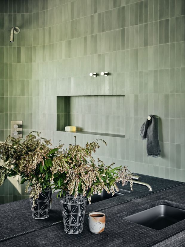 A refined bathroom with pale green vertically arranged wall tiles, a recessed open niche, wall-mounted chrome tapware, and a dark stone vanity with an integrated basin. Large leafy flowering branches in patterned vessels and a small ceramic cup sit on the counter, adding an organic decorative element.