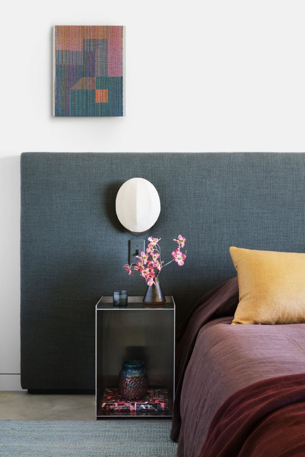 A styled bedroom vignette with a tall charcoal-blue upholstered headboard spanning the wall behind the bed. The bed is dressed in deep plum and burgundy bedding with a mustard yellow cushion. A small dark bedside table holds a vase of pink flowering branches and a glass, while a white oval wall light is mounted above. A colourful geometric textile artwork hangs on the white wall, and a muted blue-grey rug covers part of the floor.