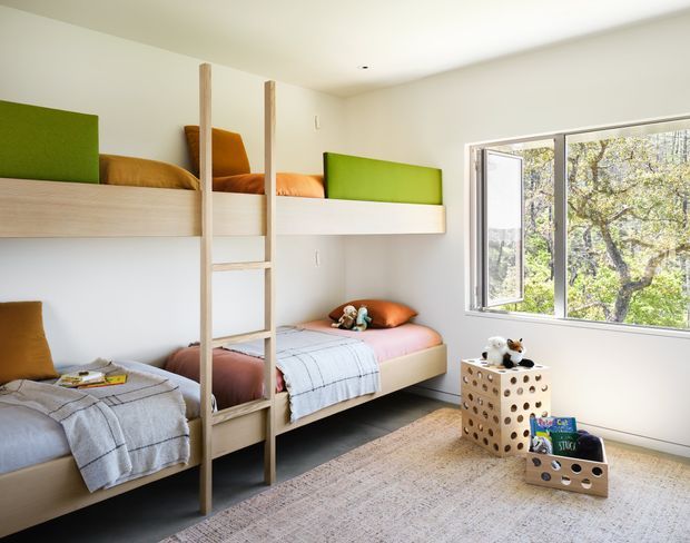 Bright, compact children's bedroom with built-in timber bunk beds arranged along two walls. The beds have colourful green, orange, and pink bedding, a central timber ladder, and soft neutral textiles. A large opening window brings in abundant natural light and views of trees. The room includes a pale rug and perforated wooden storage boxes with toys, creating a playful, functional shared sleeping space.