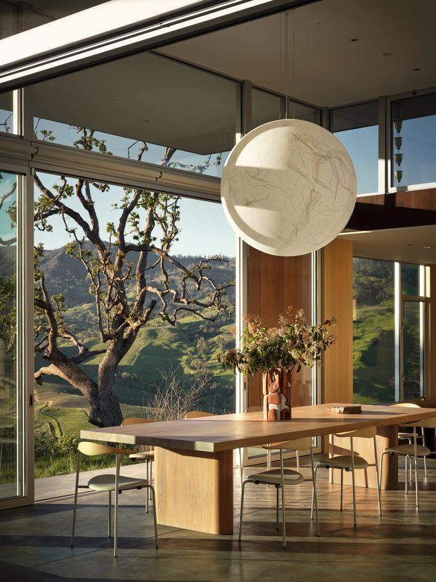 A contemporary dining area with a long light-wood dining table and integrated central pedestal, surrounded by minimalist chairs. Full-height glazing frames a dramatic green rural valley and mature trees, while a large sculptural spherical pendant light hangs above the table. Warm timber wall panelling, dark-framed glazing, polished concrete flooring, and abundant natural light create a refined indoor-outdoor connection.