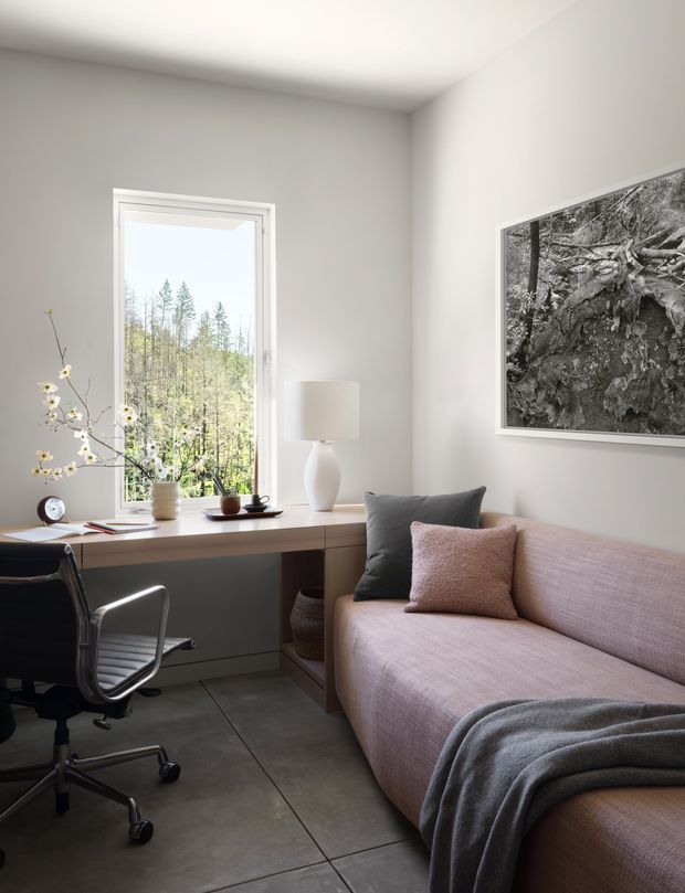 A compact, bright home office and sitting area with a built-in desk beneath a tall window, black office chair, small table lamp, and a blush-pink upholstered sofa with cushions and a grey throw. White walls and ceiling, large grey concrete-look floor tiles, minimalist furnishings, and a framed black-and-white nature photograph create a calm, refined interior. Woodland is visible through the window.