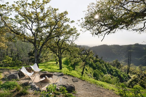 A sunlit outdoor seating area on a rocky, landscaped hillside, with several light-coloured lounge chairs arranged around a low circular fire pit. Mature trees frame expansive views across wooded rolling hills.