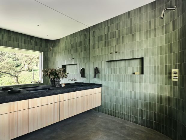 Contemporary bathroom with a long floating light-wood vanity and dark stone benchtop, set against full-height vertically arranged green-grey tiles. The room includes multiple basins, wall-mounted fixtures, recessed shelving, a wall-mounted shower outlet, and a large window overlooking greenery. Dark flooring and a restrained palette create a refined, spa-like atmosphere.