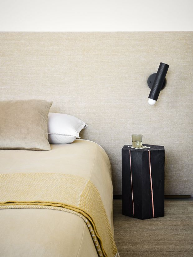 Close-up of a softly styled bedroom corner featuring a cream upholstered bed with beige and white pillows, a textured mustard throw, a black sculptural bedside pedestal, and a black wall-mounted reading light against a textured neutral wall.