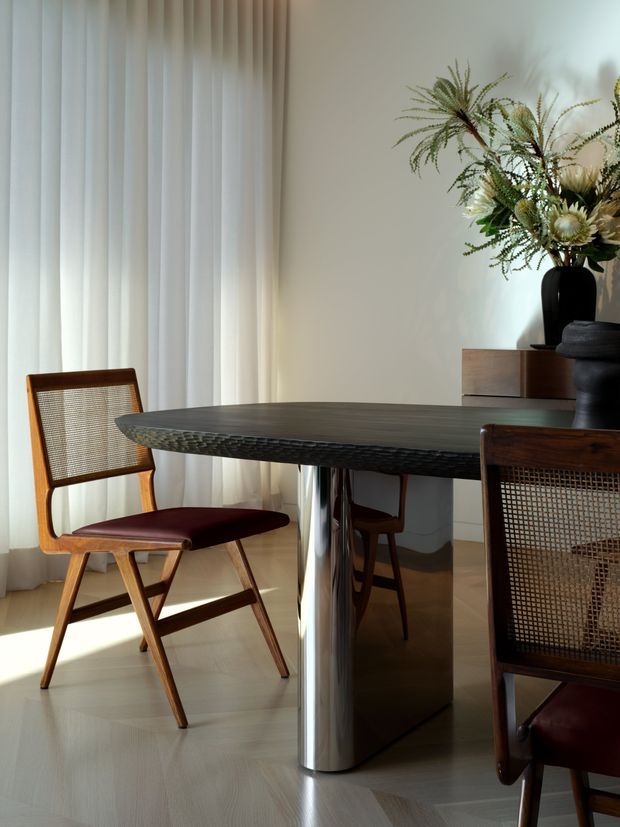 A refined dining area with a dark, sculptural oval dining table supported by a polished cylindrical metal pedestal. Cane-backed wooden dining chairs with deep burgundy seats surround the table. Sheer full-height curtains filter soft daylight, while a low wooden sideboard and large floral arrangement add warmth and texture. The room has pale timber flooring and neutral walls, creating an elegant contemporary setting.