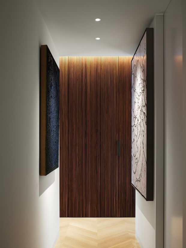 A narrow, refined residential hallway or entry corridor with white walls, recessed ceiling lights, and a warm herringbone timber floor. The focal point is a dark timber slatted feature wall and concealed door at the end, flanked by large framed artworks or decorative panels. The space has a minimalist, contemporary feel with strong vertical detailing and warm ambient lighting.