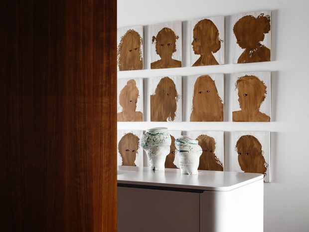 A refined interior vignette viewed beside a dark timber-paneled opening. A white built-in display wall holds a grid of portrait artworks depicting stylized brown-haired figures, with two white sculptural vessels arranged on a curved white console below. The space is minimalist and gallery-like, with strong contrast between the dark wood panel and bright white cabinetry and walls.