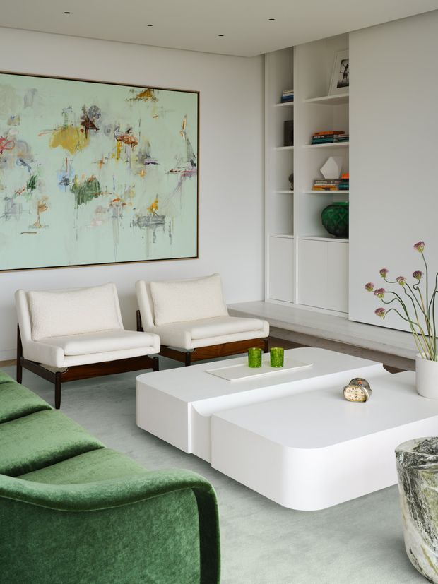 A bright, refined contemporary living room with a large abstract artwork, two cream upholstered lounge chairs, a deep green sofa, and sculptural white rounded coffee tables. Built-in white shelving displays books and decorative objects, while a pale grey floor and white walls create a clean, gallery-like setting with subtle recessed ceiling lights.
