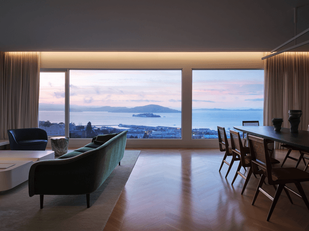 A sophisticated open-plan living and dining room with expansive floor-to-ceiling glazing overlooking a coastal bay and islands. The room features a dark ceiling, warm concealed lighting, full-height curtains, polished timber flooring, a large area rug, a sculptural green sofa, contemporary lounge seating, and a long dark dining table with wooden chairs. The panoramic outlook and reflective floor create an elegant, tranquil atmosphere.