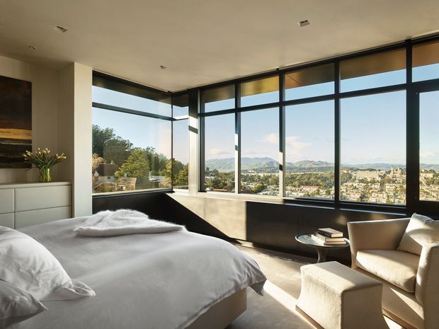 Bright contemporary bedroom with expansive floor-to-ceiling corner glazing, dark window frames, a low-profile bed dressed in white bedding, light upholstered lounge chairs, and a small round side table. The windows provide a wide elevated view over a city and surrounding hills. The room has a restrained neutral palette and abundant natural light.
