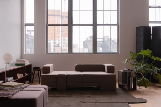 A contemporary living room in a loft-like urban interior, furnished with a low dark brown modular sofa, matching sectional seating in the foreground, a large muted area rug, and a minimalist open shelving unit. Tall black-framed industrial-style grid windows provide abundant daylight and views of city buildings. Indoor plants, books, and sculptural objects add warmth and texture.