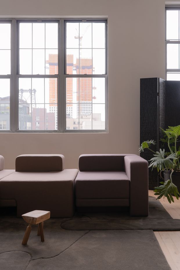 A softly lit contemporary living room with large industrial-style gridded windows overlooking an urban setting. The room features a low modular dark brown sectional sofa, a small rustic timber stool, a large dark organic-shaped rug, a black textured room divider, and leafy indoor plants. The palette is warm and muted with taupe walls, dark furnishings, and pale timber flooring.