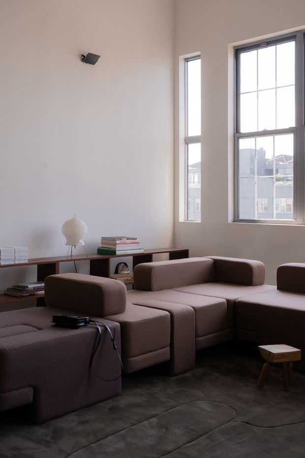 A refined living room with a large modular taupe sectional sofa arranged around a low seating area. The room features tall, multi-pane industrial-style windows with dark frames, pale plaster walls, dark low-profile shelving, stacked books, a sculptural table lamp, and a dark plush rug. Natural daylight creates a calm, understated atmosphere.