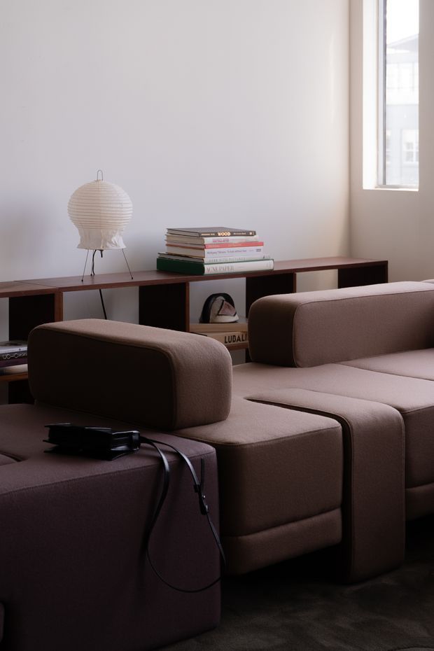 A softly lit living room featuring a large modular upholstered sectional sofa in muted taupe-brown, with rounded cushions and integrated ottoman-like sections. A long dark timber open shelving console runs along the wall behind it, displaying stacked books and a sculptural table lamp with a pleated paper shade. A small high-set window brings in daylight, while the overall scheme is restrained and contemporary.