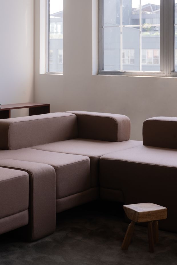 A softly lit, minimalist living room with a large modular upholstered sectional sofa in muted taupe, a dark carpeted floor, and a small rustic timber stool. Tall industrial-style metal-framed windows bring in daylight, with a simple narrow timber console along the wall.