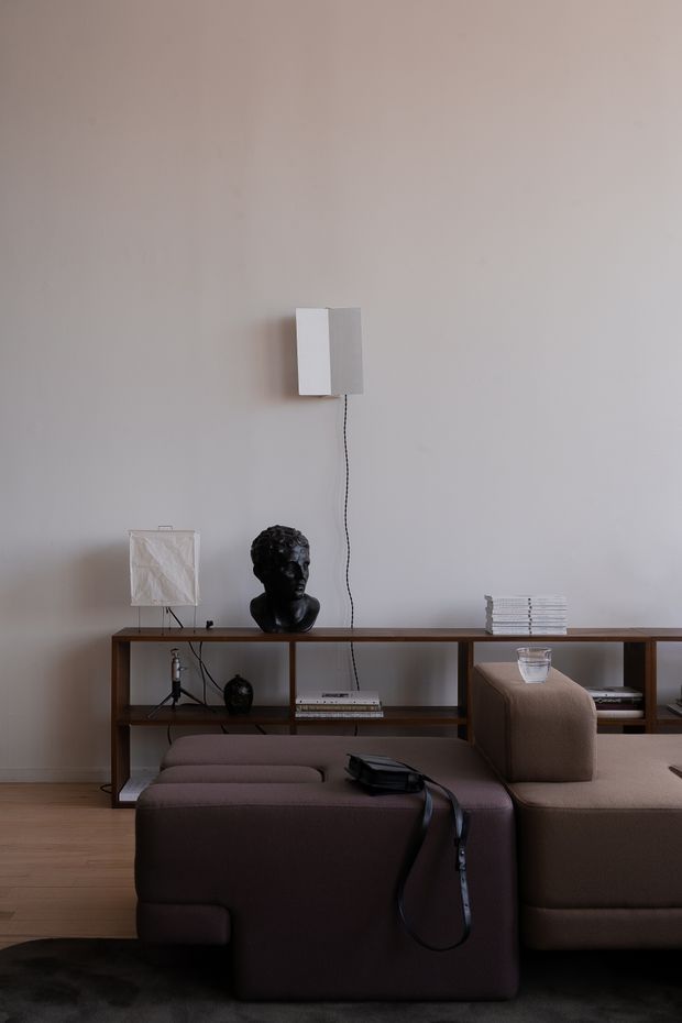 A restrained, warm-toned living room with a low modular brown sectional sofa in the foreground, a dark wood open shelving console behind it, and a minimalist wall-mounted rectangular light. The shelving displays a black sculptural bust, small decorative objects, books, stacked magazines, and a small glass vessel. Pale neutral walls, timber flooring, and subdued lighting create a calm contemporary atmosphere.