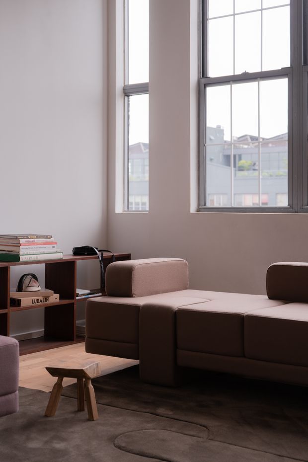 A softly lit living room with tall industrial-style multi-pane windows, pale walls, timber flooring, and a dark brown modular sofa. A low wooden open shelving unit displays books and small objects, while a simple wooden stool and large muted rug complete the understated interior.