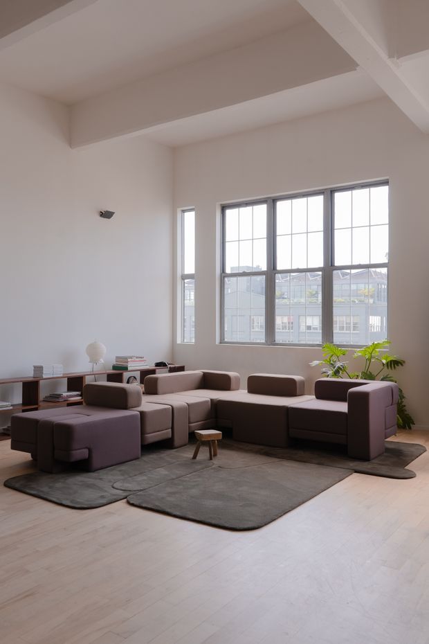 A bright, spacious living room with a high ceiling, exposed structural beams, tall multi-pane industrial-style windows, pale timber flooring, and a modular taupe-brown sectional sofa arranged over layered dark grey rugs. A low timber shelving unit, small stool, books, table lamp, and indoor plants furnish the room. The space has a clean contemporary loft-like character with abundant natural light.