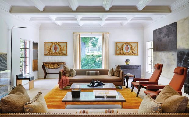 A grand, brightly lit formal living room with ornate traditional detailing and a contemporary furniture arrangement. The room features a coffered and beamed ceiling with elaborate crown moulding, white walls, large divided-light windows, a warm timber floor, and a dramatic dark stone or textured feature wall. Two sofas face a central glass-topped coffee table, complemented by orange leather lounge chairs, a carved console table, framed artwork, and sculptural decor.