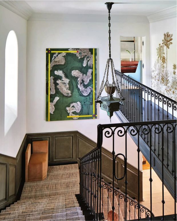 A refined interior stairway with a split-level landing, ornate black wrought-iron balustrades, patterned carpeted stairs, dark timber wall panelling, white plaster walls with decorative cornices, and a traditional hanging lantern. A large abstract artwork and a glimpse of an adjacent room are visible.