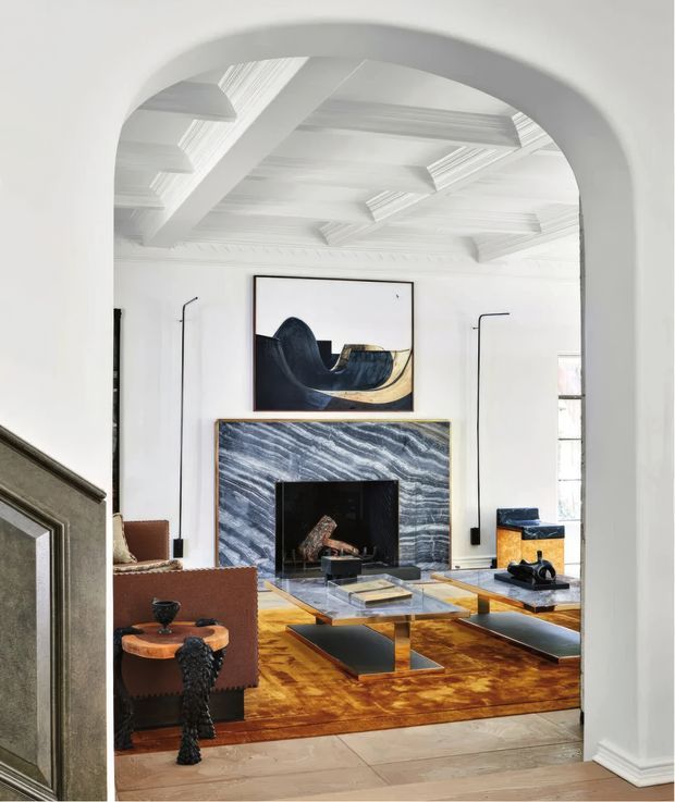 A refined living room viewed through a broad arched opening. The room features an ornate white coffered and beamed ceiling, white walls, a dramatic blue-grey veined stone fireplace surround, dark contemporary furniture, a large patterned rust-toned rug, sculptural lighting, and abstract artwork. The space combines classical architectural detailing with eclectic contemporary furnishings.