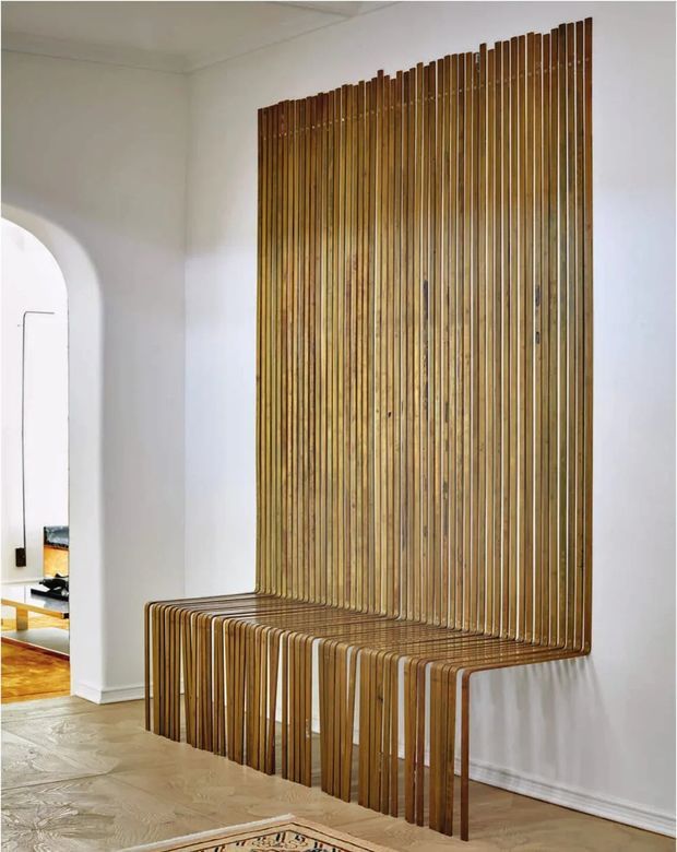 Interior view of a bright white room featuring a sculptural built-in timber slat bench that rises into a full-height vertical feature wall. The continuous curved slats create seating and backrest elements, with an arched opening leading to another room on the left. Light timber flooring and minimal white walls give the space a clean, contemporary character.