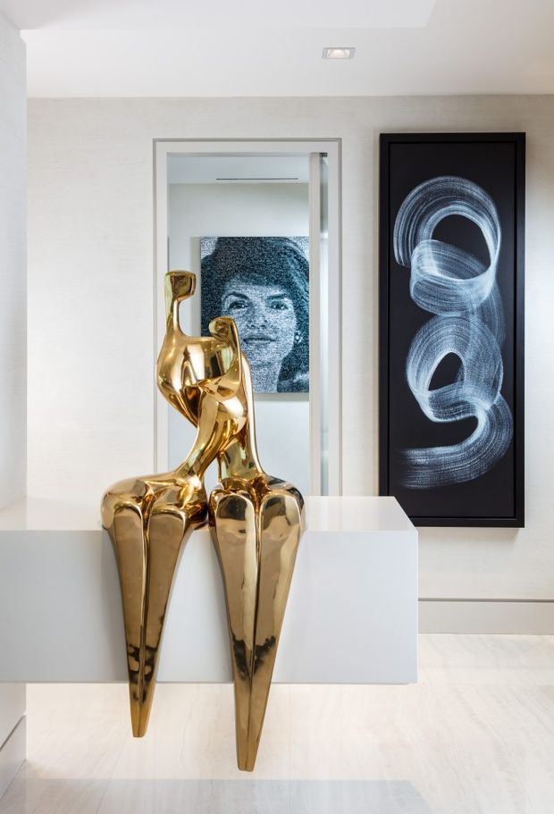 A bright, refined interior vignette featuring a white built-in console or plinth with a highly polished gold sculptural artwork in the foreground. A recessed opening or doorway behind it contains another monochrome portrait artwork, while a tall black-framed abstract artwork hangs on the textured white wall to the right. Pale flooring, white walls, and recessed ceiling lighting create a clean gallery-like atmosphere.