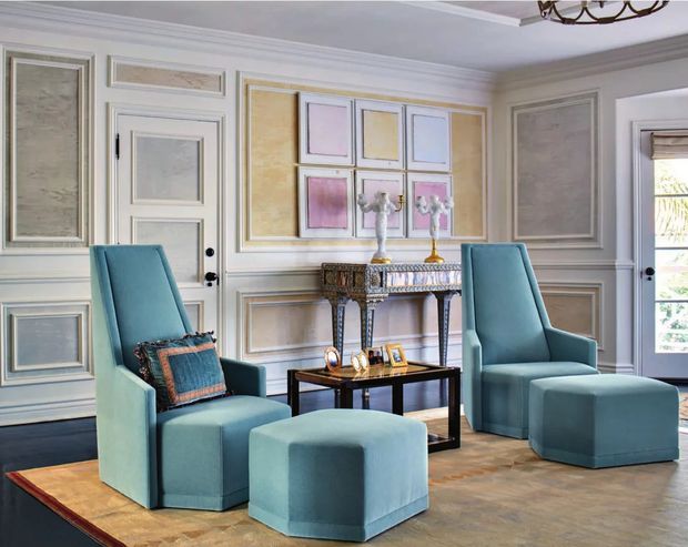 Elegant formal sitting room with pale blue upholstered armchairs and matching ottoman, a dark wood and glass coffee table, and an ornate console table. The room features elaborate painted wall panelling, decorative mouldings, framed artwork, a parquet-style timber floor, and a traditional chandelier, creating a refined classical interior.