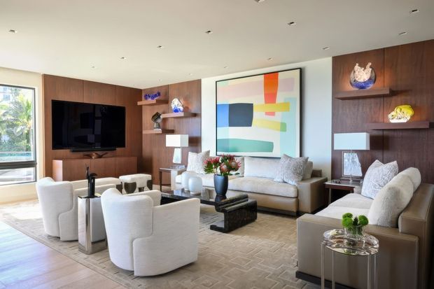 Bright, refined living room with a large sectional sofa, sculptural upholstered armchairs, a glass-topped coffee table, and built-in dark timber wall panelling. The room features a wall-mounted television, open display shelving, contemporary abstract artwork, recessed ceiling lights, and expansive glazing with a leafy view outside.